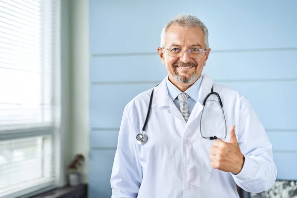 Smiling medical senior doctor with a stethoscope. On a blue background. Medic shows thumb raised up. The concept of humanitys victory over disease