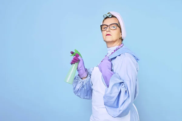 Portrait of old cleaning woman in apron with dust cleaning brush isolated on blue background. special uniform and professional equipment for cleaning