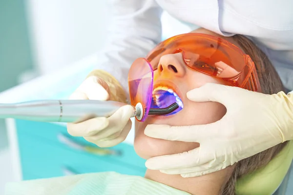 Girl child at the doctor. Dentist places a filling on a tooth with dental polymerization lamp in oral cavity. over clinic background — Stock Photo, Image