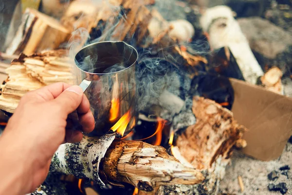 Man traveler hands holding cup of tea near the fire outdoors. Hiker drinking tea from mug at camp. Coffee cooked over a campfire on the nature. Adventure, travel, tourism and camping concept.