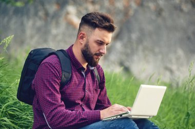 Sakallı adam ağacın altındaki parkta dizüstü bilgisayarla çalışıyor ve gülümsüyor. Happy Freelancer çimlerin üzerinde oturuyor ve uygulama ya da web sitesi kullanıyor. Uzaktan izolasyon modunda çalış