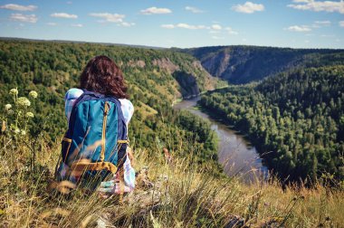 Sırt çantalı mutlu genç bir kadın uçurumun kenarında oturur ve aşağıdaki vadiye bakar.