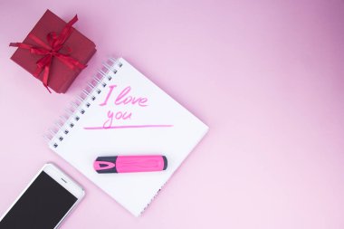 Kırmızı kurdeleli bir hediye. Akıllı telefon hediyenin yanında. Defterdeki yazı. Seni seviyorum.