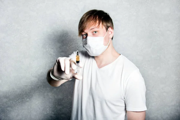 A man in a medical mask holds a syringe with medicine. A graduate student holding a syringe in a mask and medical gown isolated on a white background. Concept of medicine.