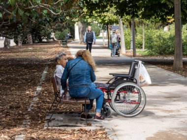 Madrid, İspanya, 9 Ekim 2019. Parktaki bankta yaşlı bir bayanla birlikte bakım işçisi. Tekerlekli sandalye yanında..
