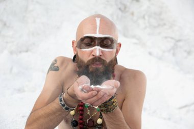 İlkel makyajlı ve boncuklu genç adam beyaz kayaların üzerinde ellerinde pudrayla büyüler yapıyor.