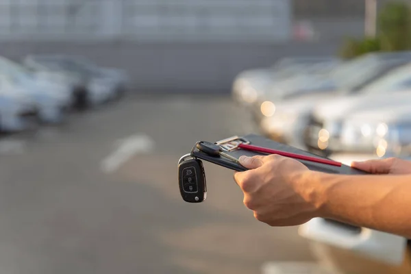 Händerna på en bilhandlare som håller nycklar och urklipp till en kund, mot bakgrund av de fordon parkerade. — Stockfoto