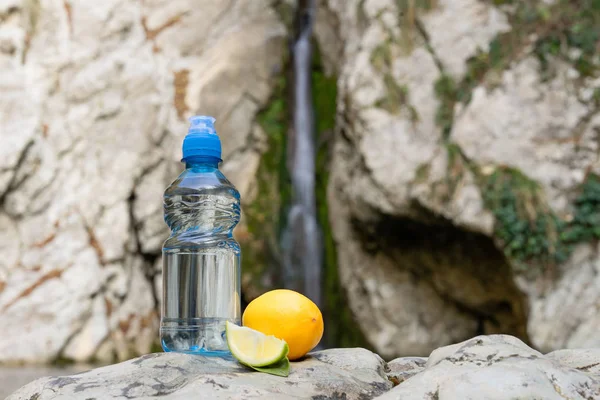 Bir dağ nehri ve şelalenin arka planında limon tadı olan çocuklar için şişe suyu. — Stok fotoğraf