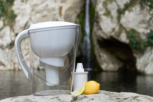Empty water filter jug with lemon, stands on a stone near a waterfall