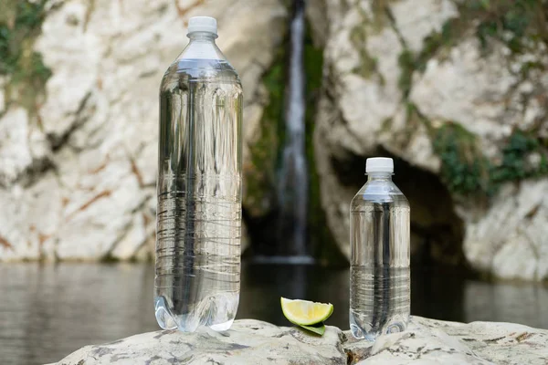 Dos botellas de agua potable con un limón en el fondo de un lago de montaña y cascada. Agua mineral todavía — Foto de Stock