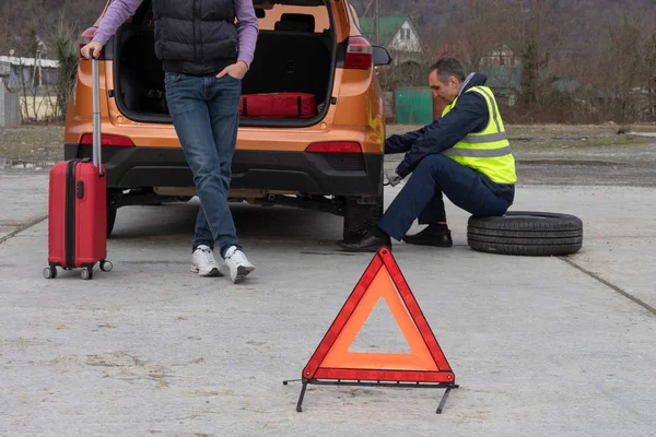 Unge man får hjälp med reservhjulsbyte. Slaghjul på vägen under körning. Mekanikern är klädd i en reflekterande väst som ersätter däck. Nödstoppskylt inställd framför. — Stockfoto