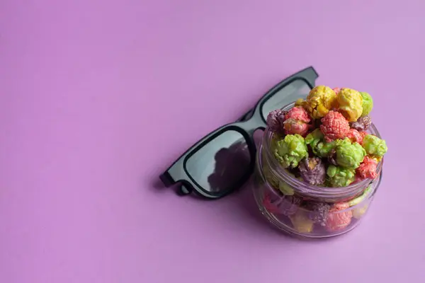 Multicolored fruit flavored popcorn in glass cup with cinema 3D glasses on pink background. Candy coated popcorn.