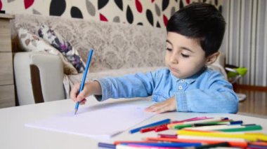 Masada oturan ve beyaz kağıda renkli kalemler çizen sevimli bir çocuk..