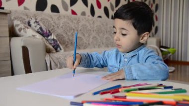Masada oturan ve beyaz kağıda renkli kalemler çizen sevimli bir çocuk..