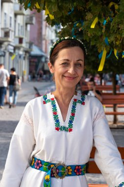 Chernivtsi, Ukrayna, 25.08.2019: Eski şehir merkezinin yaya sokağındaki ağaç ve ahşap bankın yanında duran beyaz elbiseli mutlu esmer kadın