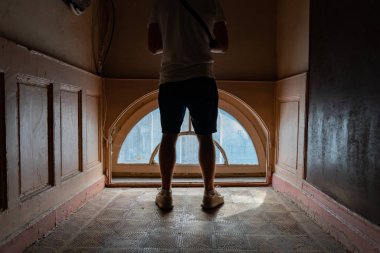 Şortlu genç adam eski ahşap antika pencerenin yanında kameranın karşısında duruyor.