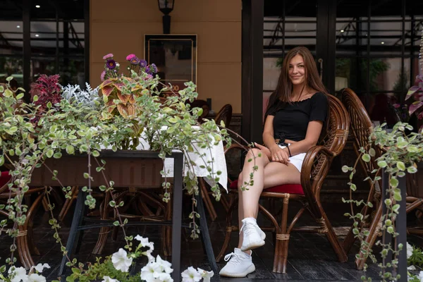 Chica joven morena feliz en camiseta negra sentada en una terraza de una cafetería decorada con flores — Foto de Stock