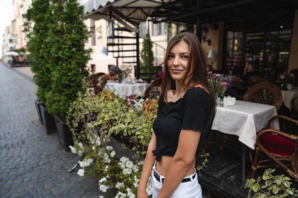 검은 티셔츠를 입은 행복 한 갈색 소녀가 꽃으로 장식된 카페 거리 테라스에 앉아 카메라를 보고 있다. — 스톡 사진