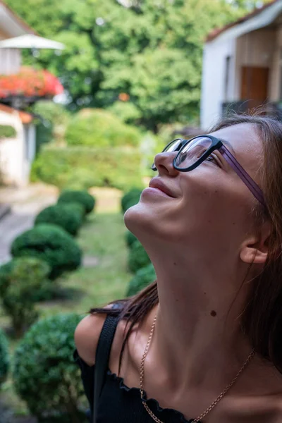 Close up portrait of beutiful brunette girl in glasses that lookes up, green patio on the background — ストック写真