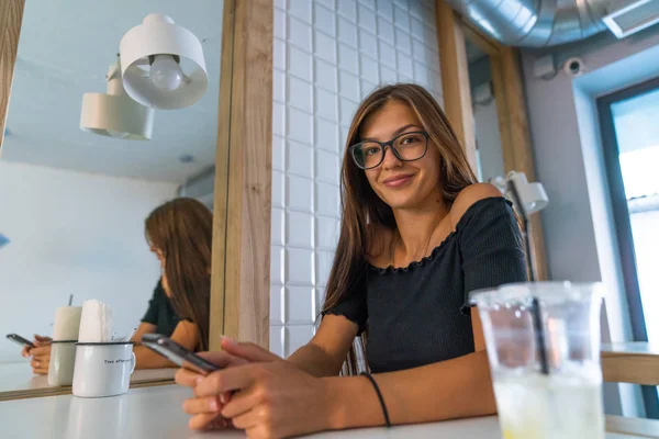 Happy cute brunette girl with loose hair in glasses sitting at a table in a cafe, holds her phone in hands and looking at the camera — Stock Photo, Image