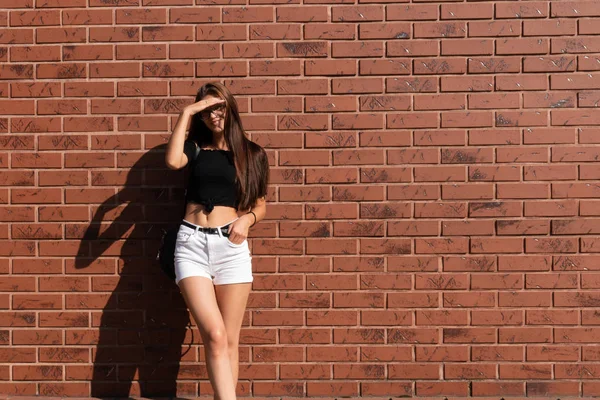 Young brunette girl with loose hair, dressed in white shorts and black top standing near a red brick wall and covers her eyes with her hand from the sun