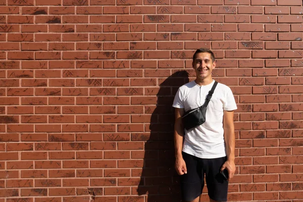 Jovem sorridente feliz em t-shirt branca e com ombro preto de volta stand perto da parede de tijolo vermelho e olhando para a câmera — Fotografia de Stock