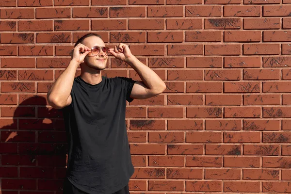 Atractivo joven en camiseta negra de pie cerca de la pared de ladrillo rojo sosteniendo gafas de sol con las manos — Foto de Stock