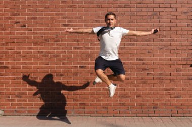 Beyaz tişörtlü genç adam elleri çapraz atlıyor, kollarını ardına kadar açıyor ve kameraya bakıyor, arka planda kırmızı tuğla duvar var.