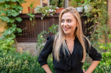 Gülümseyen, gevşek saçlı mutlu sarışın kız elleri belinde ve başka tarafa bakıyor, arka planda yeşil saksılarla süslenmiş eski bir balkon.