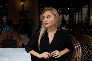 Chernivtsi, Ukrayna, 25.08.2019: Siyah elbiseli çekici sarışın kız, rahat bir sokak kafesinde masada oturuyor.