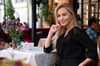 Chernivtsi, Ukrayna, 25.08.2019: Gevşek saçlı ve doğal makyajlı çekici sarışın Gil, siyah elbiseli, çiçeklerle süslenmiş rahat bir sokak kafesinde oturuyor.