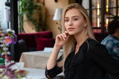 Sarışın, bol saçlı, doğal makyajlı, siyah elbiseli, rahat bir sokak kafesinde çiçeklerle süslenmiş bir masada oturan zarif bir bayan.