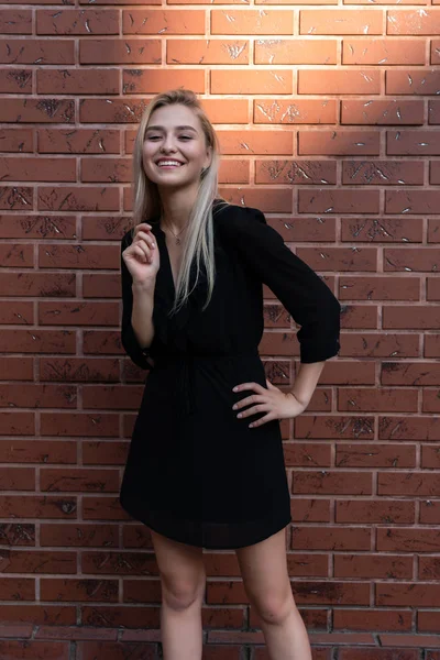 Joyeux sourire fille blonde joyeuse en robe noire tient une main sur sa taille l'autre près de son visage, mur de briques rouges sur le fond — Photo