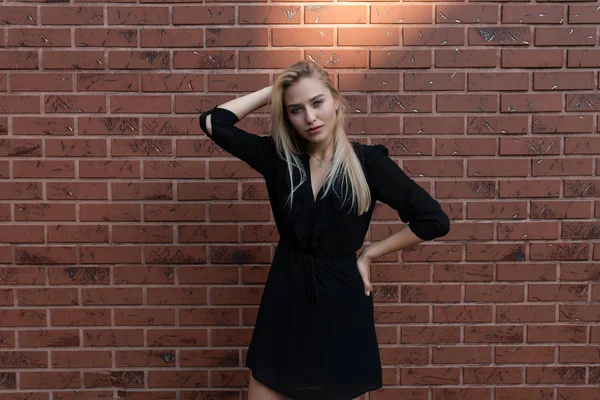 Modische blonde Frau in schwarzem Kleid hält eine Hand auf der Taille, die andere auf dem Kopf und blickt in die Kamera, rote Backsteinmauer im Hintergrund — Stockfoto