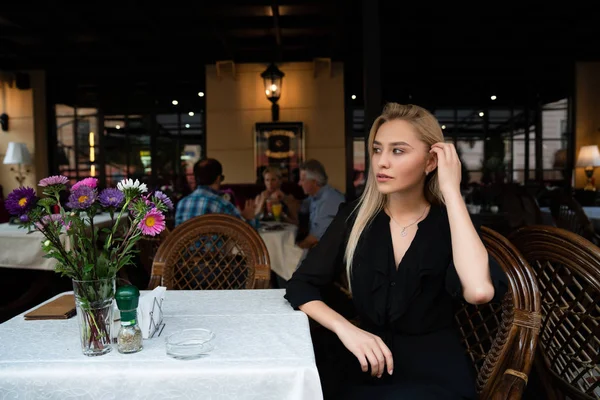Chernivtsi, Ucrânia, 25.08.2019: Menina loira misteriosa em vestido preto sentada em uma mesa em um aconchegante café de rua decorado com flores e endireita o cabelo com a mão — Fotografia de Stock
