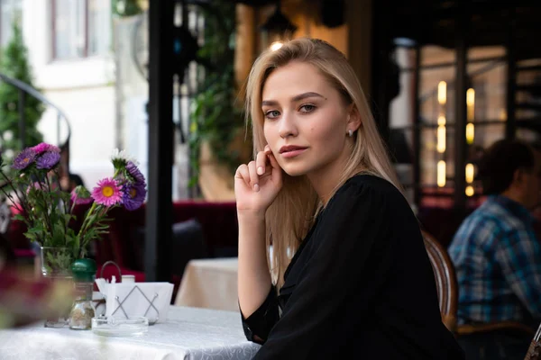 Tajemnicza blondynka w czarnej sukience siedzi przy stole w przytulnej ulicznej kawiarni ozdobionej kwiatami i prostuje włosy dłonią — Zdjęcie stockowe