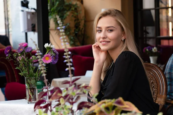 Elegancka pani z blond luźne włosy i naturalny makijaż, ubrana w czarną sukienkę siedzi przy stole w przytulnej ulicznej kawiarni ozdobionej kwiatami — Zdjęcie stockowe