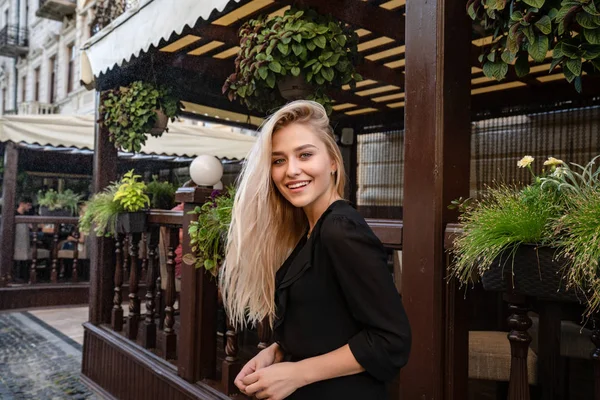 Szczęśliwa uśmiechnięta blondynka z naturalnym makijażem i luźne włosy, ubrana w czarną sukienkę stojącą w pobliżu letniego tarasu — Zdjęcie stockowe