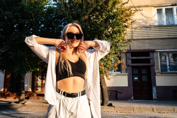 Menina loira elegante em óculos de sol pretos vestidos com shorts brancos e camisa de pé entre a rua com as mãos levantadas — Fotografia de Stock