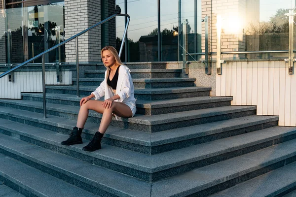 하얀 셔츠와 검은 부츠를 신고 계단에 앉아 있는 예쁜 금발 소녀의 야외 생활 방식 사진 — 스톡 사진