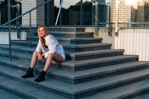 Привлекательная улыбающаяся блондинка в белой рубашке и черных сапогах сидит на лестнице — стоковое фото
