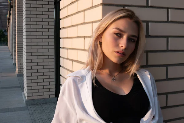 Sensual blonde girl with natural makeup dressed in black top leaned on the white brick wall and looks at the camera — ストック写真