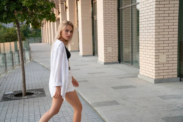 Seriamente menina loira com cabelo solto, vestida com camisa branca e botas pretas caminha ao longo do edifício moderno — Fotografia de Stock