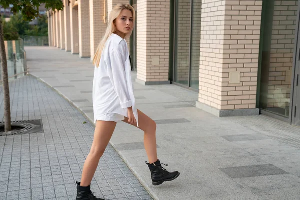 Vážně blondýnka s volnými vlasy, oblečená v bílé košili a černé boty procházky podél moderní budovy — Stock fotografie