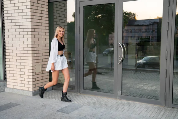 Menina loira sorridente feliz em camisa branca e botas pretas caminha ao longo do edifício moderno — Fotografia de Stock