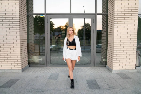 Модная стильная блондинка в белой рубашке и черных сапогах гуляет по современному зданию — стоковое фото