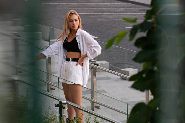 Moda chica rubia en camisa blanca y pantalones cortos de pie cerca de la barandilla de cristal —  Fotos de Stock