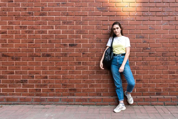 Attractive serious young brunette girl in blue jeans and yellow t-shirt, with black shoulder bag isolated over red brick wall — Stock Photo, Image