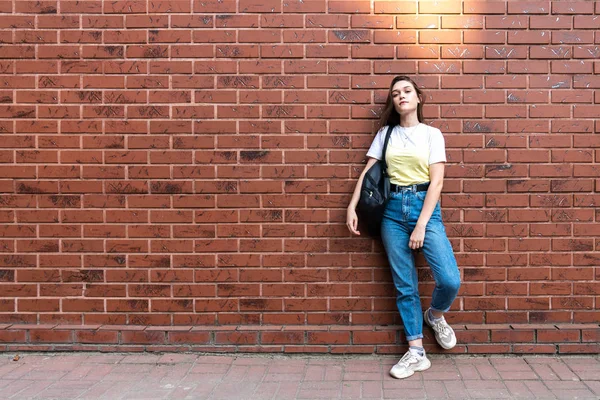 Atractiva joven morena seria en jeans azules y camiseta amarilla, con bolso negro aislado sobre pared de ladrillo rojo — Foto de Stock