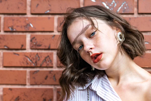 Close up portrait of the beautiful brunette girl with short wavy hair, red lips and freckles dressed in white striped shirt isolated over red brick wall — 스톡 사진